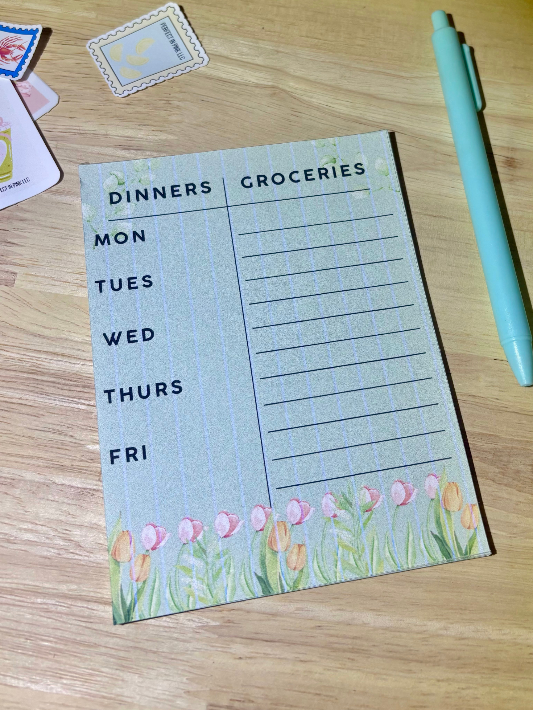 A weekly planner notepad with a 'Dinners' and 'Groceries' table, decorated with pink tulips at the bottom, shown on a wooden desk next to a mint green pen.